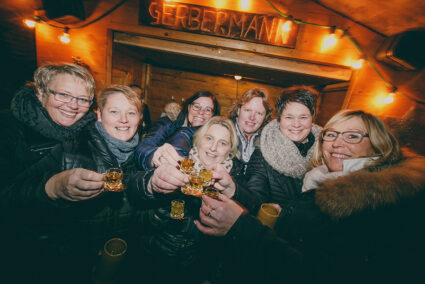 Eine Gruppe Frauen steht vor einer Weihnachtsmarkthütte und stößt mit einem Schnaps an