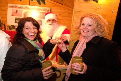 Zwei Frauen stehen an einer Weihnachtsmarkthütte und trinken einen Schnaps mit einem verkleideten Weihnachtsmarkt