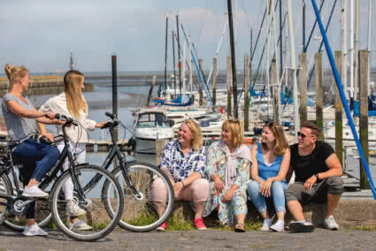 Drei Frauen und ein Mann sitzen auf einer kleinen Mauer an einem Yachthafen und gucken zwei Frauen mit ihren Fahrrädern an die neben ihnen stehen