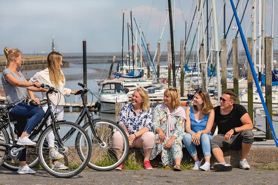 Drei Frauen und ein Mann sitzen auf einer kleinen Mauer an einem Yachthafen und gucken zwei Frauen mit ihren Fahrrädern an die neben ihnen stehen